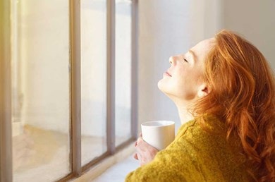 Eine Frau sitzt mit geschlossenen Augen und einer Tasse vor einem geschlossenen Fenster und lässt die Sonne in ihr Gesicht scheinen