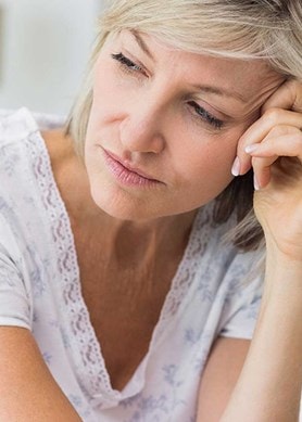 Eine Person in einem weißen Spitzenoberteil sitzt nachdenklich mit der Hand an der Stirn auf einem Bett vor neutralem Hintergrund.