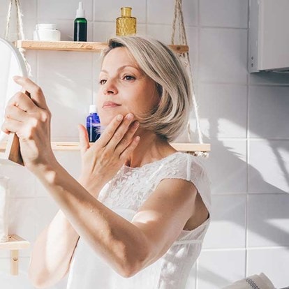 Une personne tient un miroir dans une salle de bain lumineuse, entourée de plantes et de produits de soin disposés sur une étagère en bois.