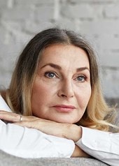 Person mit blonden Haaren lehnt entspannt auf einem grauen Sofa, im Hintergrund eine helle Backsteinwand. Kleidung in weiß gehalten.