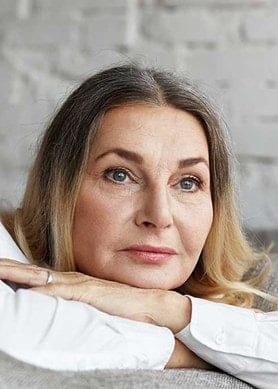 Person mit blonden Haaren lehnt entspannt auf einem grauen Sofa, im Hintergrund eine helle Backsteinwand. Kleidung in weiß gehalten.