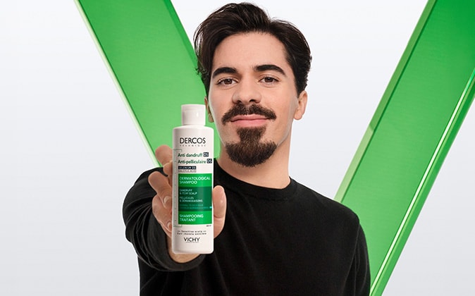 Person hält eine Flasche Vichy Dercos Anti-Schuppen-Shampoo für empfindliche Kopfhaut vor grünem Hintergrund in die Kamera