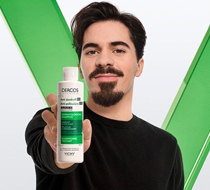 Person hält eine Flasche Vichy Dercos Anti-Schuppen-Shampoo für empfindliche Kopfhaut vor grünem Hintergrund in die Kamera