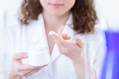Une femme teste une crème blanche provenant d'un pot sur son doigt, avec en arrière-plan un laboratoire bleu et des détails flous.