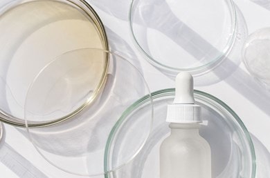 Flacons en verre avec pipette, entourés de boîtes de Pétri transparentes et de récipients en verre sur fond blanc, éclairés par une lumière vive.