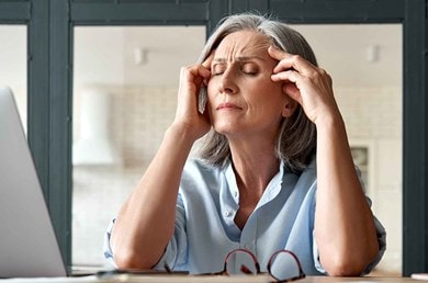 Une femme aux cheveux gris a retiré ses lunettes et se tient la tête, le visage crispé par la douleur.