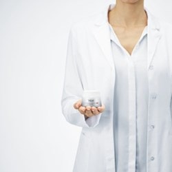 Une femme au visage non visible, portant une chemise et une blouse blanches, tient un pot de crème Vichy dans sa main