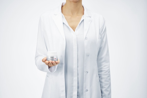 Une femme au visage non visible, portant une chemise et une blouse blanches,  tient un pot de crème Vichy dans sa main
