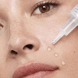 Gros plan sur une femme aux yeux bruns avec trois gouttes de sérum sur la joue et un flacon de sérum ouvert en face d'elle
