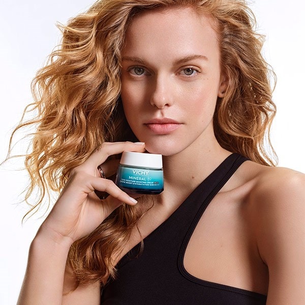 Une femme tient le pot de crème MINÉRAL 89 100H Hydratation Boost dans sa main. Son visage est visible en arrière-plan