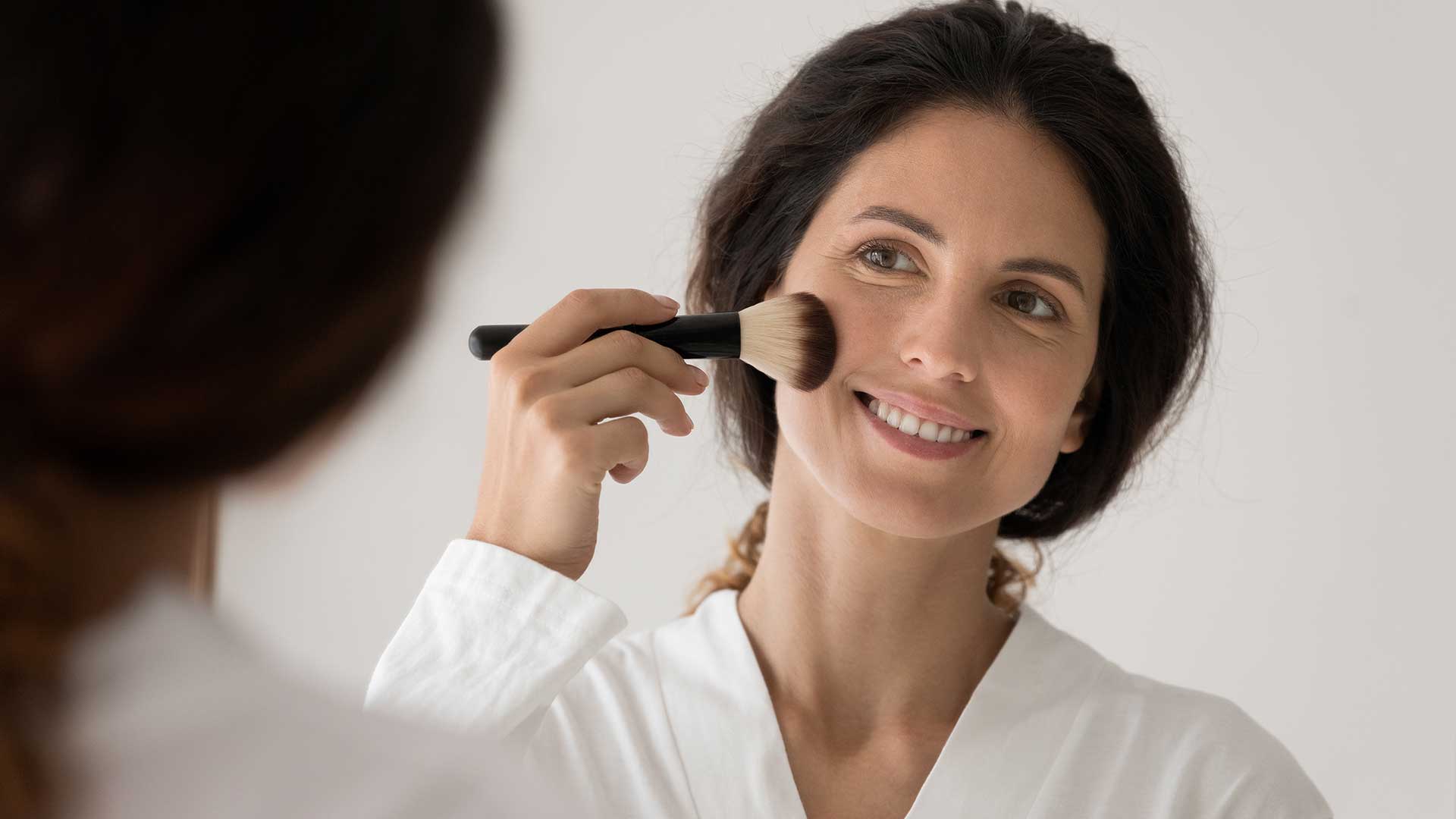 Lächelnde junge Frau mit glatter Gesichtshaut hält einen Kosmetikpinsel an ihre Wange 