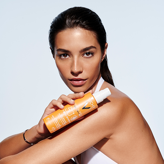 Junge Frau mit einem Lächeln im gesicht hält die CAPITAL SOLEIL Cell Protect Sonnenspray LSF 50 von Vichy in der Hand 